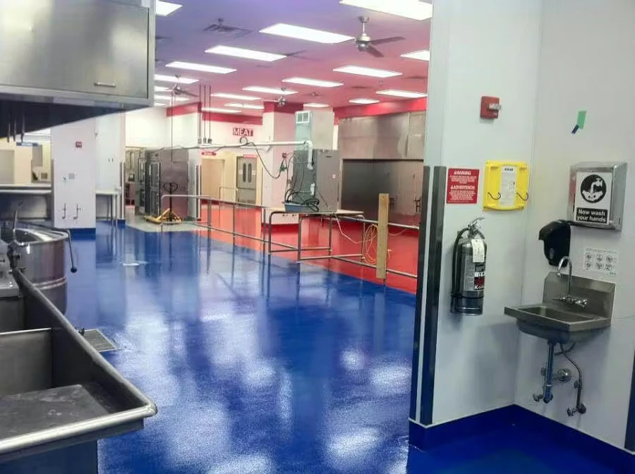Commercial kitchen or food-processing room with stainless equipment and a glossy blue epoxy floor, leading toward a red-floored area; a wall-mounted sink, soap dispenser, and fire extinguisher are in the foreground.