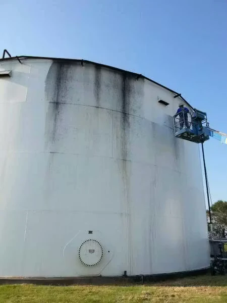 A large white cylindrical storage tank with dark vertical streaks of grime running down from the top edge, while a worker on a lift platform paints or cleans the upper side against a clear blue sky.