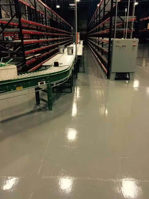 Warehouse interior with tall pallet racking aisles and a green conveyor line in the foreground; the floor is coated in shiny gray epoxy, reflecting overhead lights, with an electrical control box near the conveyor.
