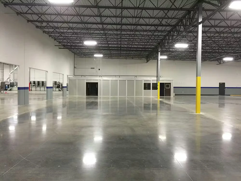 Empty industrial warehouse interior with a high steel-truss ceiling, bright overhead lights, and a smooth polished concrete floor reflecting the lights; several support columns include yellow safety paint, and a glass-partitioned office area sits along the back wall.