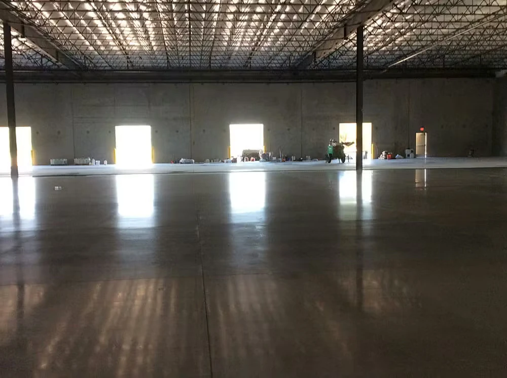 Wide view inside a large warehouse with a high truss ceiling and a glossy, polished concrete floor reflecting light from several open loading-bay doors along the far wall; small equipment and supplies sit near the openings.