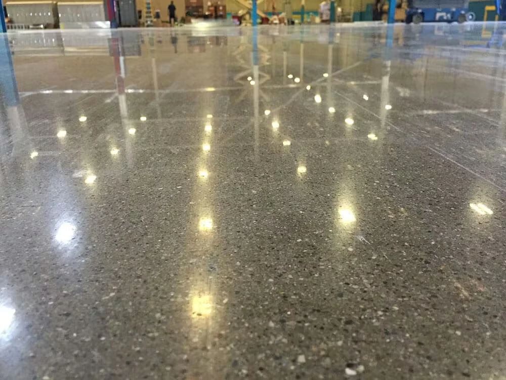 Close-up of a glossy polished concrete floor in a warehouse or factory, with bright overhead lights reflecting across the smooth surface; background shows blue support columns and industrial equipment out of focus.