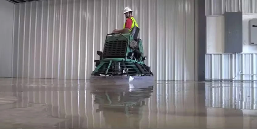 Worker in a hard hat and safety vest rides a power trowel across a glossy concrete floor inside a metal-walled industrial building, with the machine and lights reflected on the polished surface.