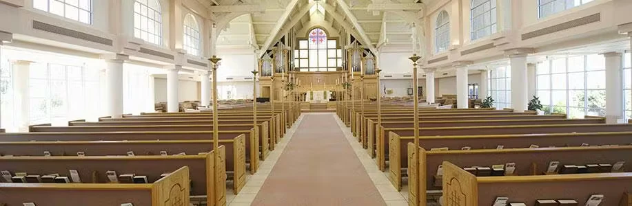Wide view down the center aisle of a bright church sanctuary with rows of wooden pews, white columns, large arched windows, and an altar area at the far end with a stained-glass window above it.
