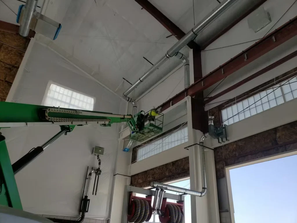 Interior of a tall industrial bay with white walls and exposed red steel beams; a worker in a green boom lift basket is raised near the ceiling, working alongside large silver HVAC ducts and vents, with high windows letting in daylight.