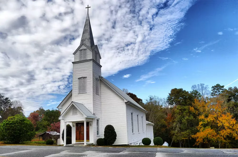 A small white wooden church with a tall steeple topped by a cross sits beside a paved road under a bright blue sky with scattered clouds, surrounded by trees with autumn foliage.