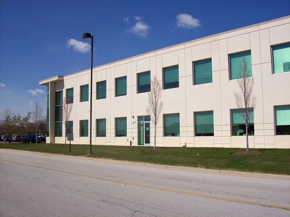 Two-story beige commercial building with teal-tinted rectangular windows and a glass corner entry, set back from a quiet street with a grass strip, small leafless trees, and a single black streetlight under a bright blue sky.