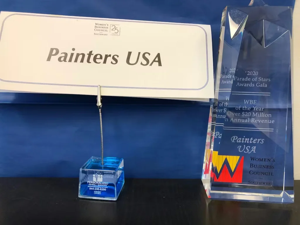 Tabletop display with a “Painters USA” name placard on a metal holder and a tall clear crystal award engraved for a 2020 Women’s Business Council Southwest “Parade of Stars” gala recognition.