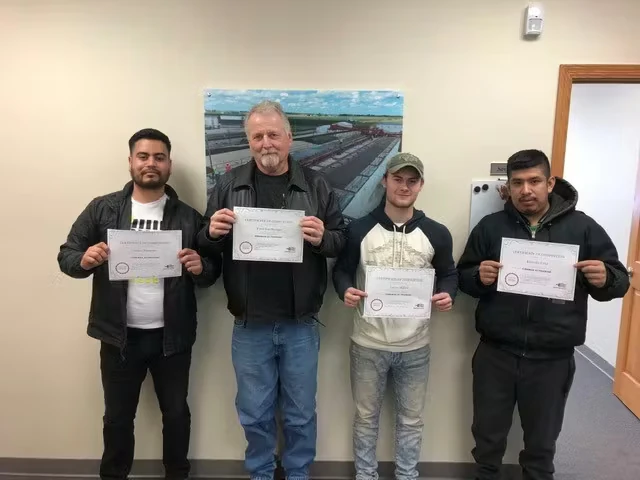 Four men stand indoors against a beige wall, each holding a certificate toward the camera; a framed photo of an industrial site hangs behind them, and a wooden doorframe is visible at the right.