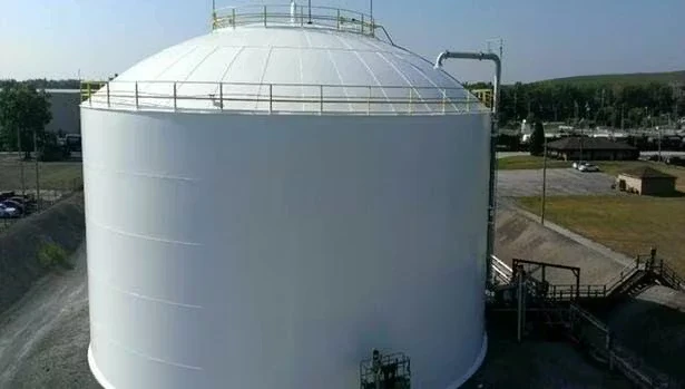 Large white cylindrical storage tank with a domed roof, perimeter railing and ladder access, viewed from above; industrial piping connects near the top, with surrounding roads and facility structures in the background.
