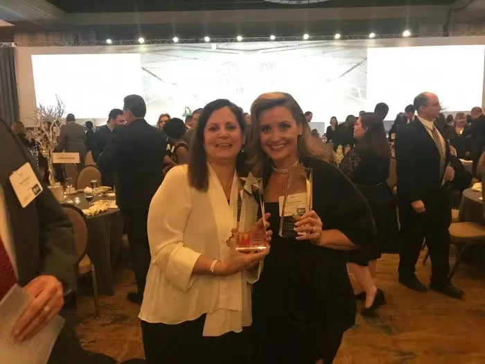 Two women stand close together at a crowded banquet-style awards event, smiling and holding clear glass trophies, with tables and formally dressed attendees in the background.