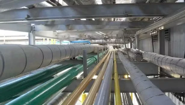 Overhead view down a long industrial pipe rack with multiple insulated gray pipes and several glossy painted pipes (green, yellow, and tan) running parallel beneath a metal roof structure.