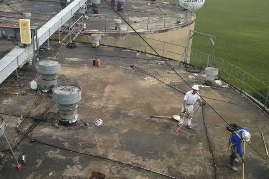 Two workers apply material across a flat industrial roof near large ventilation stacks and a metal walkway, with safety lines stretched across the surface and a grassy field visible beyond the roof edge.