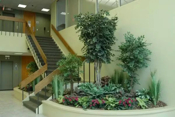Office lobby with a staircase and wood handrails beside a large curved indoor planter filled with shrubs, tall potted trees, and flowering plants under fluorescent ceiling lights.