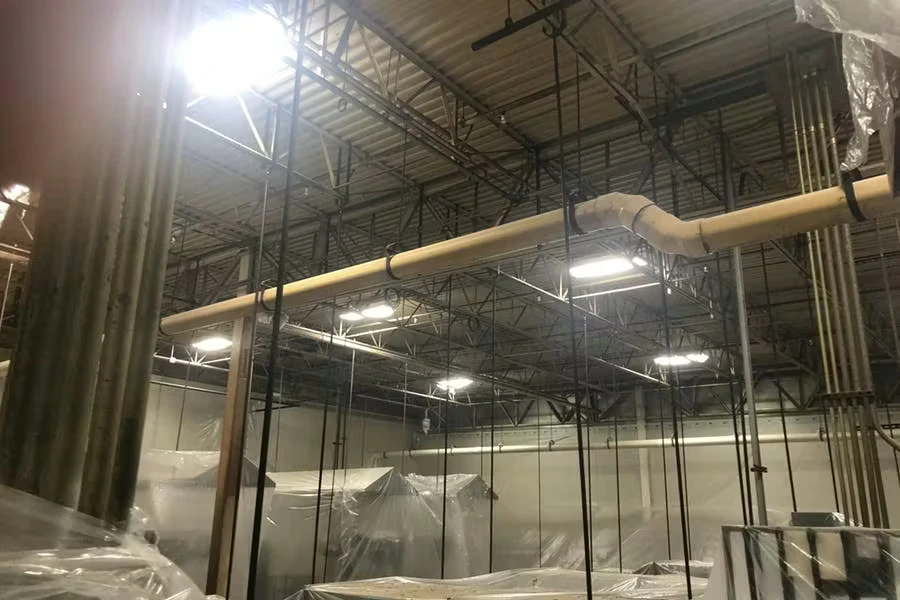 Industrial facility interior with a high open ceiling, exposed beams and hanging cables, bright overhead lights, and large pipes running across the ceiling; equipment below is partially covered with plastic sheeting.