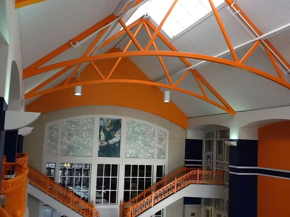 Interior atrium with a high arched ceiling and exposed orange steel trusses, orange stair railings on multiple levels, and large windows and wall panels beneath the roof.