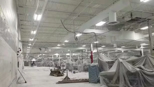 Large industrial interior with a white ceiling and rows of bright overhead lights; cables hang down in places, and many machines or work areas are covered with plastic sheeting across an expansive floor.