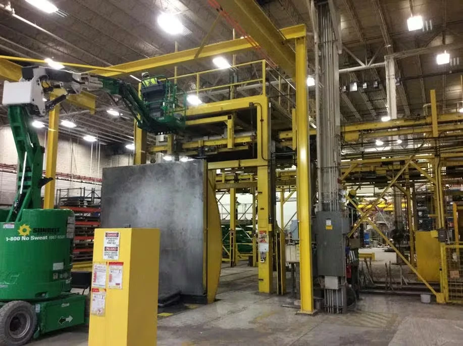 Inside an industrial facility, a worker in a green boom lift reaches an elevated yellow steel platform and catwalk above large machinery and support columns, with bright overhead lights illuminating the factory floor.