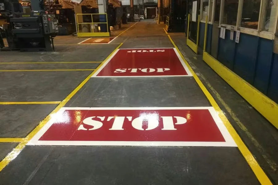 Industrial factory aisle with gray floor marked by yellow boundary lines and two large red rectangular floor stencils reading “STOP” in white, one in the foreground and one farther down the lane; blue-and-yellow machine enclosures line the right side.