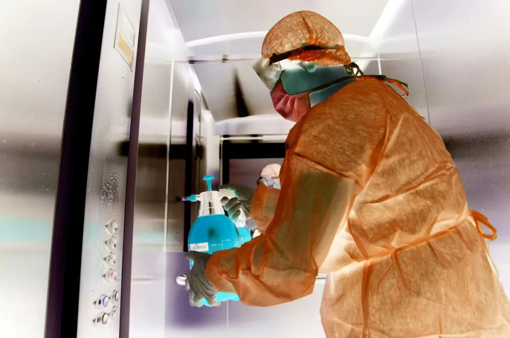 Worker in protective gear (hooded coveralls, mask, gloves, and goggles) disinfects an elevator control panel with a handheld pump sprayer; the image appears color-inverted/negative, making the sprayer look bright blue and the suit orange.