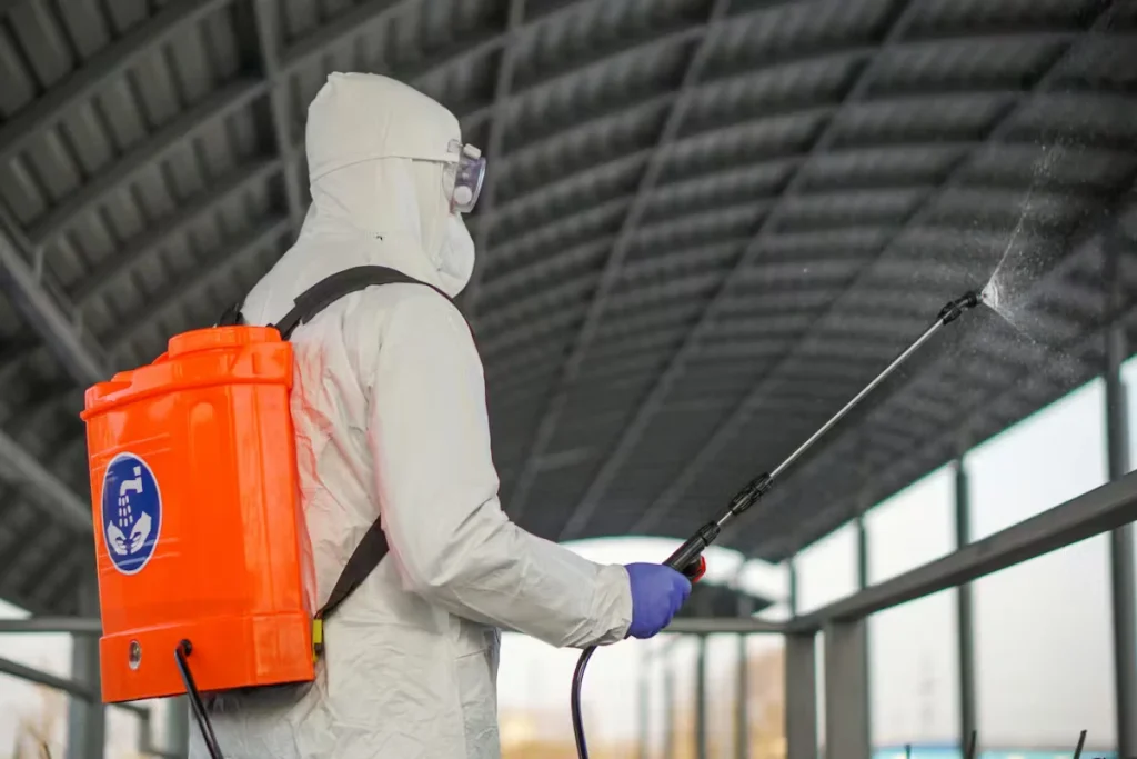 Worker in a white protective suit with goggles and blue gloves uses a long spray wand while wearing an orange backpack sprayer inside a large industrial building with a curved metal roof and tall windows.