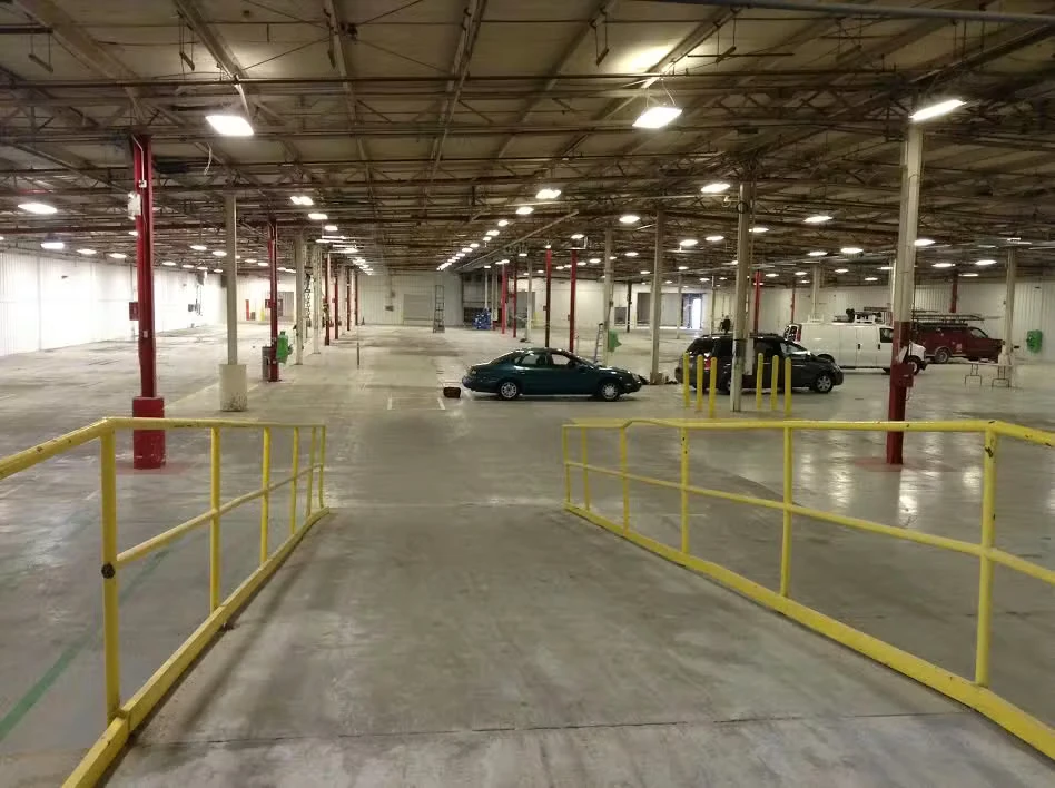 Wide view of a large warehouse interior with a polished concrete floor, rows of ceiling lights and red-and-white support columns, and yellow safety railings framing a ramp in the foreground; a few cars and equipment are parked in the open space.