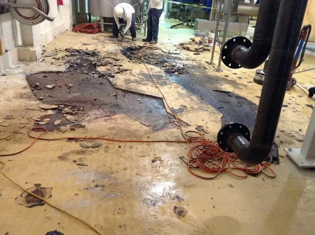 An industrial room with a damaged concrete floor where sections of old coating have been torn up into jagged piles, exposing dark patches and wet-looking areas; two workers in protective clothing work in the background while extension cords snake across the floor near large black pipes.