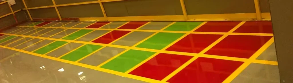 Glossy epoxy floor with a grid of bright yellow lines dividing large rectangles filled with red and green sections, next to gray areas, in an indoor industrial space with a yellow guardrail along the back wall.