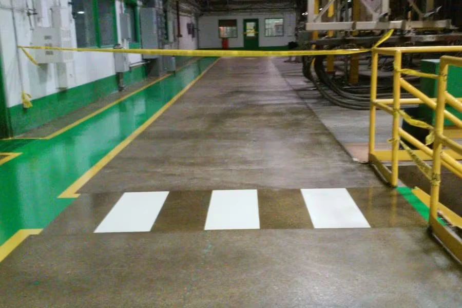 Industrial facility corridor with a concrete floor, a green-painted walkway edged with yellow lines on the left, three wide white crosswalk-style stripes in the foreground, and yellow safety railings on the right.