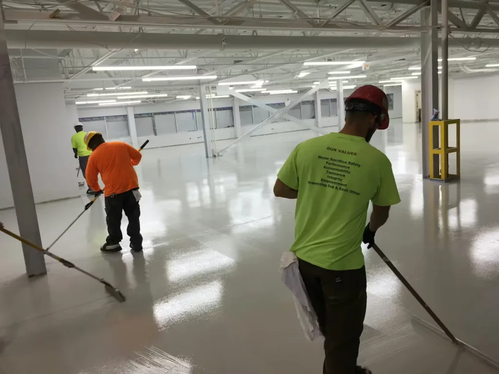 Three workers in safety gear use long-handled rollers to apply a glossy light-gray floor coating in a large, bright warehouse with white walls, exposed ceiling beams, and rows of overhead lights reflecting on the wet surface.