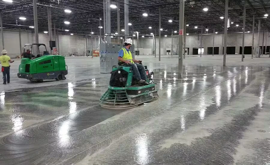 Inside a large warehouse with tall columns and bright overhead lights, a worker in a hard hat and safety vest rides a power trowel across a wet, glossy concrete floor while another green machine and worker are visible in the background.