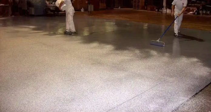 Two workers in white clothing apply a coating across a large industrial floor; one uses a long-handled squeegee while another works farther back, and the freshly coated surface looks shiny and textured under overhead lighting.