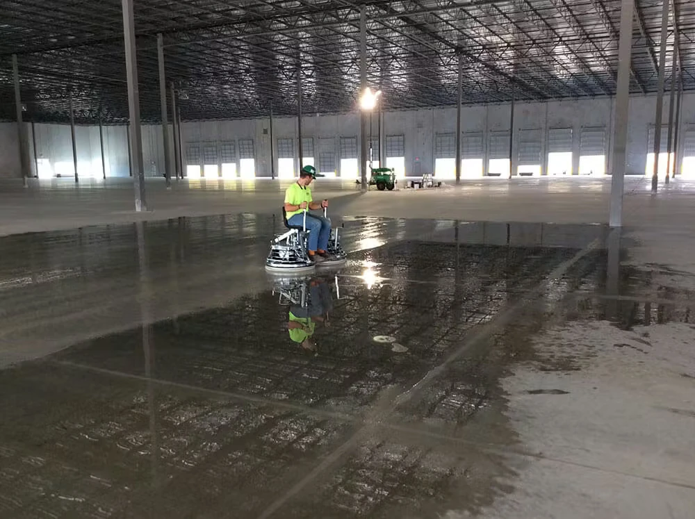 Inside a vast empty warehouse with rows of steel columns and a high grid ceiling, a worker in a neon green shirt rides a power trowel across a wet, glossy section of concrete that reflects the ceiling and lights, with bright loading bay doors lining the far wall.