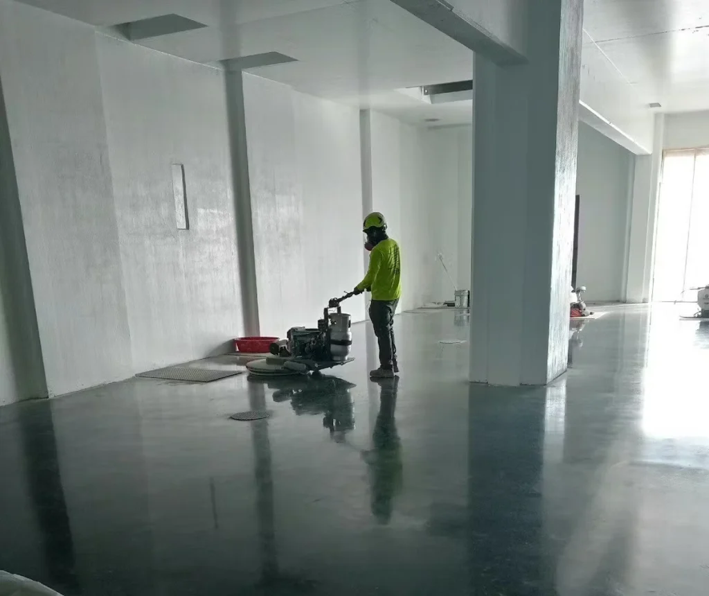 Worker in a bright safety-green shirt and respirator uses a floor burnisher/polisher on a shiny dark concrete floor inside a large white industrial room with tall columns and light from windows at the far end.