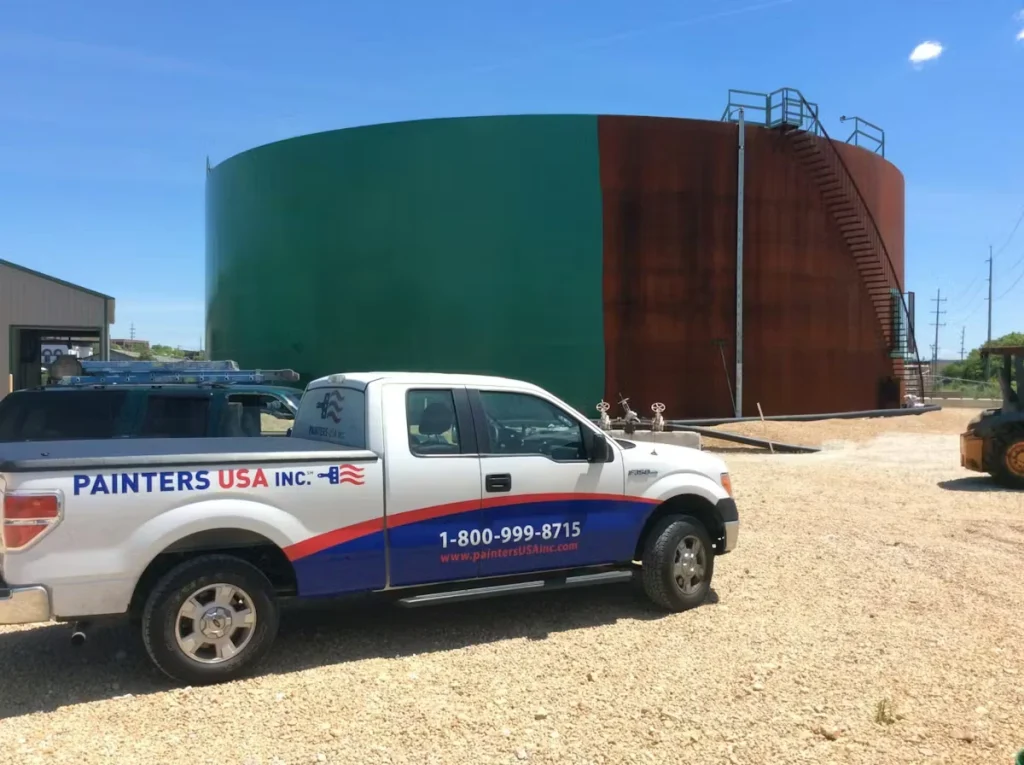 A white Painters USA pickup truck is parked on a gravel lot in front of a massive cylindrical storage tank; the tank’s left half is newly coated dark green while the right half is rust-brown, with an exterior stairway and platform on the right side under a clear blue sky.