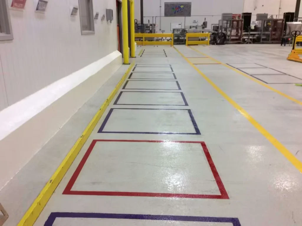 Wide industrial floor with painted line-striping—yellow lane lines and a row of outlined rectangular bays (red, purple, and dark blue) running along a wall inside a warehouse or factory.