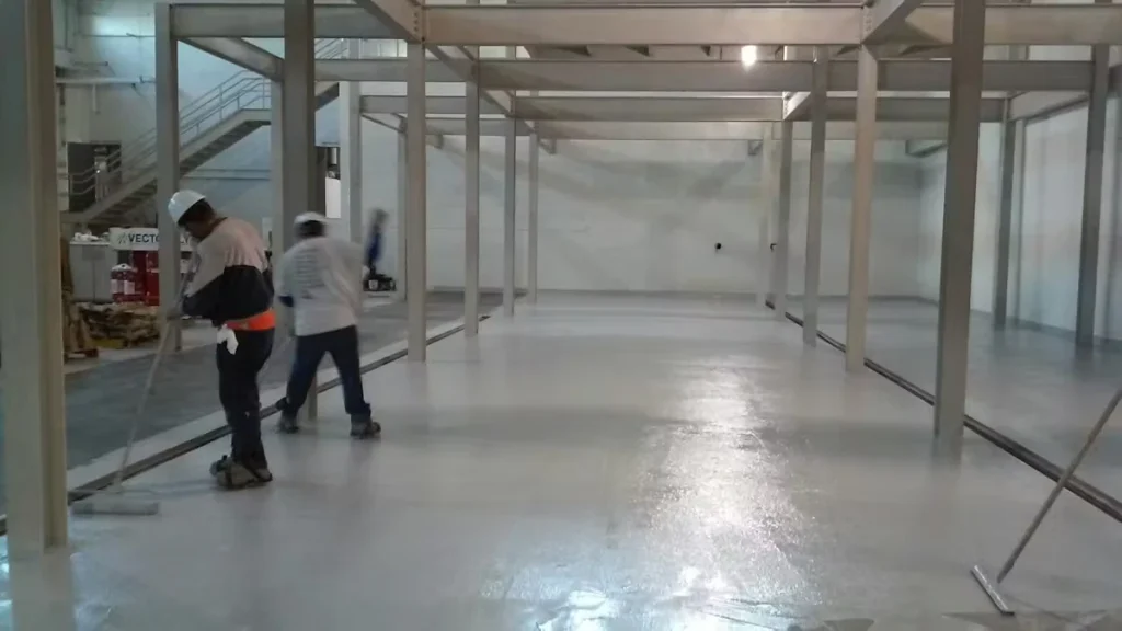 Two workers use long-handled rollers to apply a glossy light-gray epoxy floor coating inside a large industrial room with steel columns and an upper mezzanine walkway.