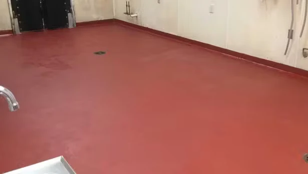 Empty utility room with cream-colored walls and a smooth red urethane-cement floor, including a small floor drain near the center and plumbing fixtures along the walls.