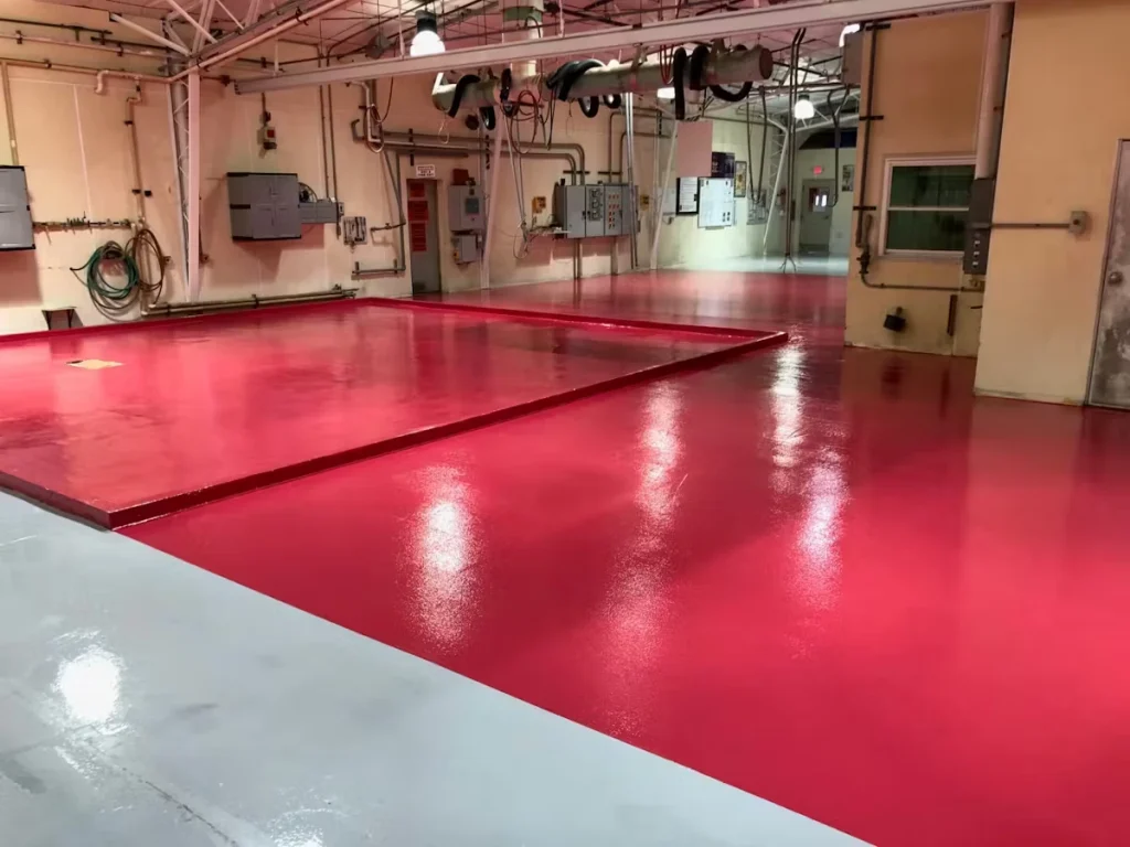 Bright red, glossy industrial floor coating inside a utility room, with overhead pipes and hanging hoses, wall-mounted electrical panels, and a raised rectangular platform section in the center reflecting ceiling lights.