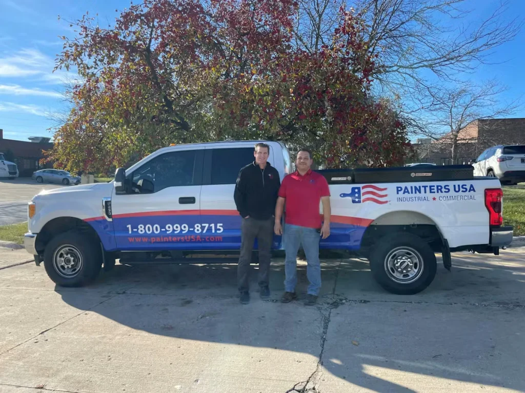 Two men stand smiling in front of a white Painters USA pickup truck with blue and red graphics and the phone number “1-800-999-8715,” parked outdoors on a sunny day with autumn trees in the background.