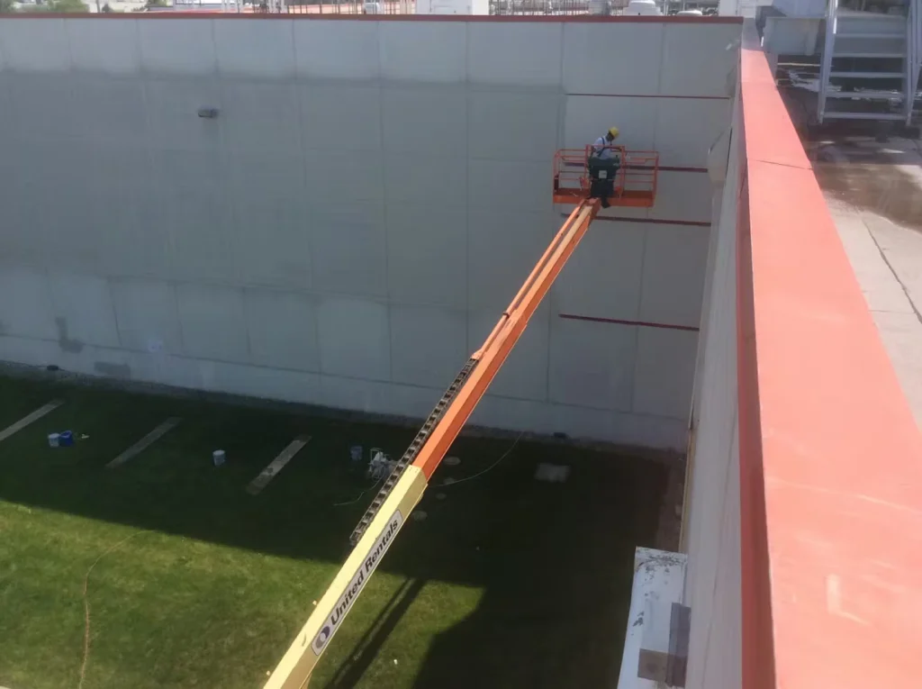 Worker in a boom lift basket near the top of a tall industrial building, painting or working on the exterior wall; the long orange lift arm extends from the ground across a grassy area, viewed from a rooftop edge.