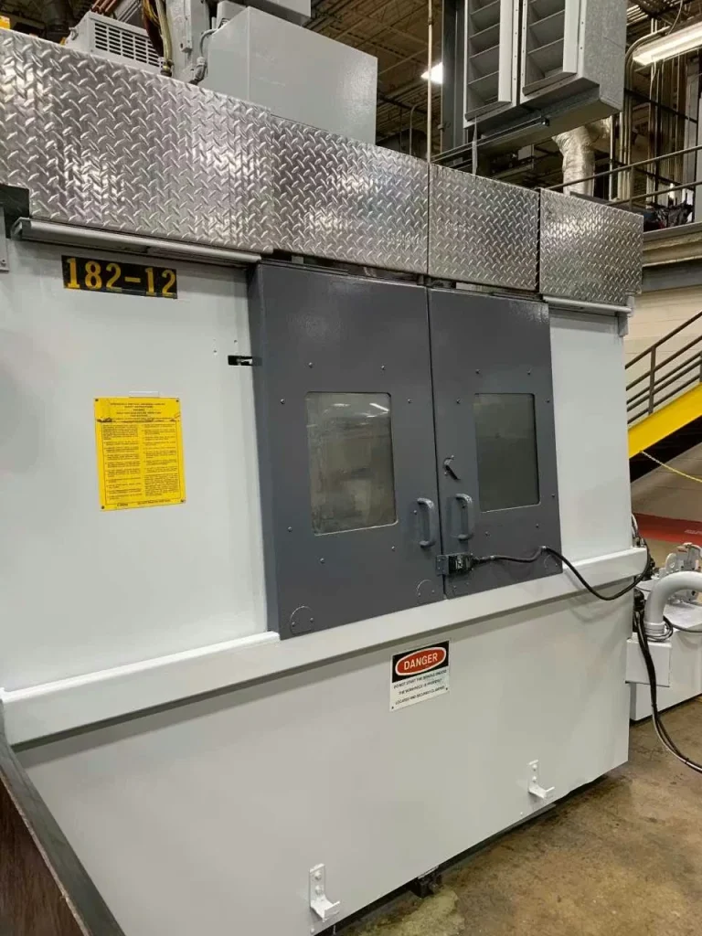 Large industrial machine in a factory, freshly painted light gray with dark gray double access doors and small viewing windows; diamond-plate metal panels run along the top, and multiple safety/ID labels are visible on the front.