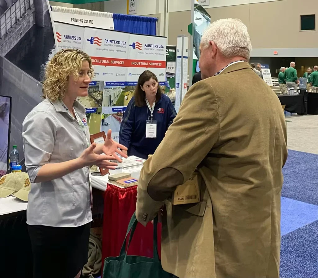 At a trade show booth for Painters USA Inc., a staff member in a gray branded shirt talks with a male attendee in a tan blazer while another staff member stands behind the table; banners for agricultural and industrial services are visible in the background.