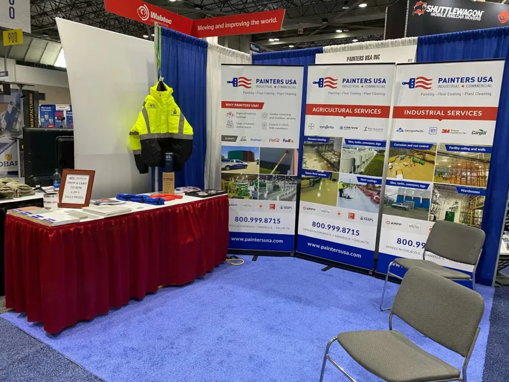 Trade show booth for Painters USA Inc. with three branded banner stands (“Agricultural Services” and “Industrial Services”), a table with brochures and giveaway items, a high-visibility jacket hanging on the side wall, and two gray chairs on blue carpet.