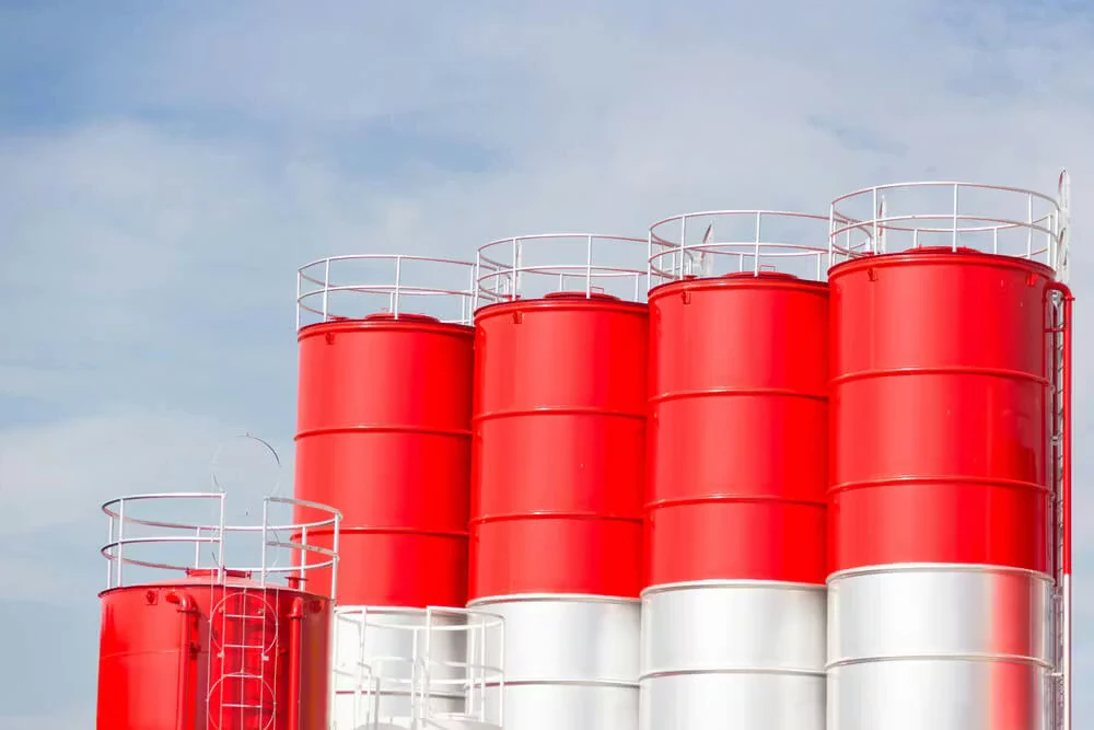 Group of large cylindrical storage tanks painted bright red with white lower sections and white safety railings along the tops, set against a pale blue sky.