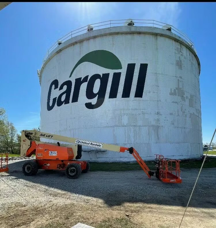 Large white cylindrical storage tank with a big “Cargill” logo painted on the side, with an orange boom lift parked in front on a gravel surface under a clear blue sky.