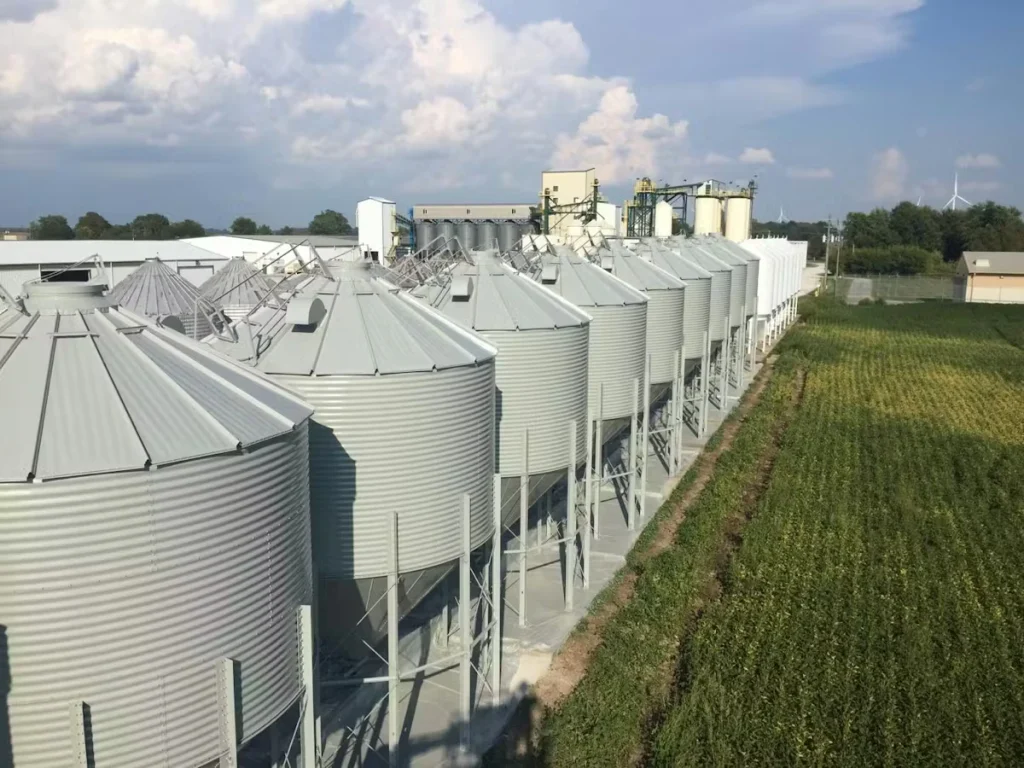 A row of large corrugated metal grain bins with conical roofs stretches alongside a green crop field, with industrial equipment and buildings in the background under a partly cloudy sky.