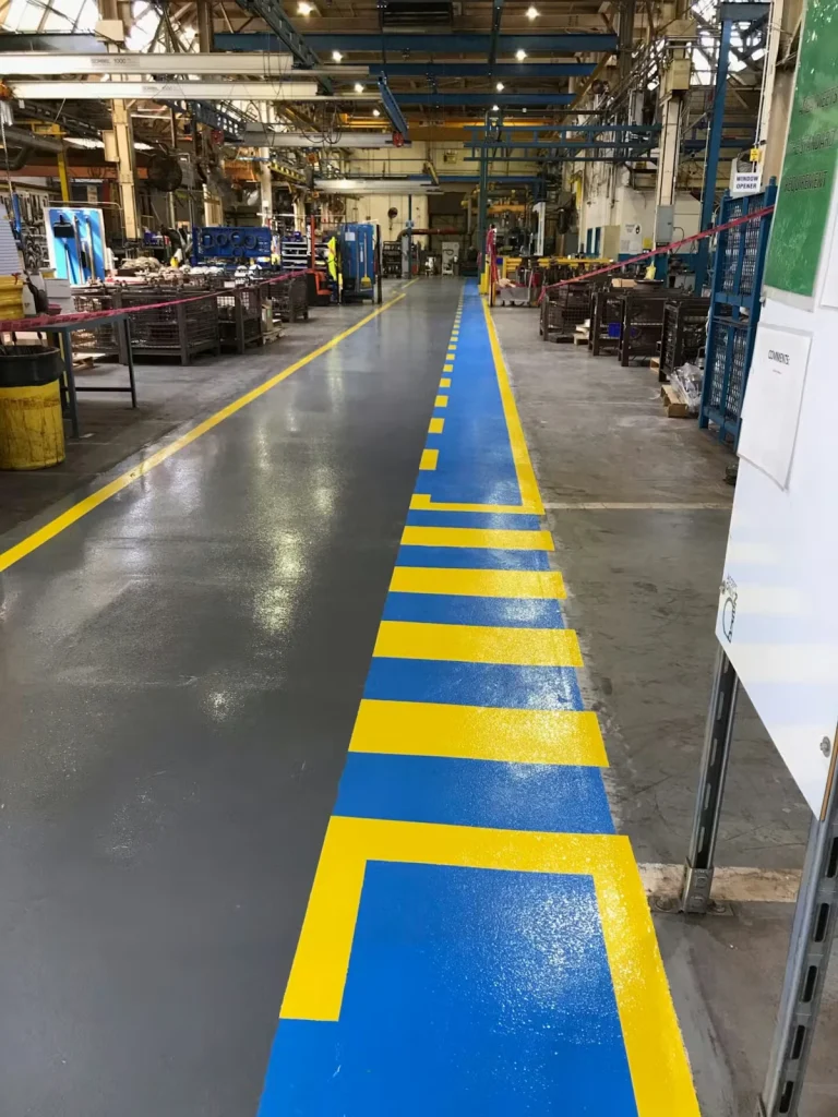 Inside a factory, a long glossy walkway runs through the center with bright blue floor paint and bold yellow striping, marking a pedestrian path alongside work areas with equipment and pallets.