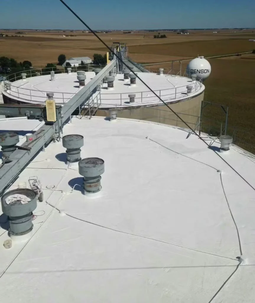 View from atop large circular storage tanks with bright white coated roofs, vents, and railings, connected by a catwalk, with flat farmland stretching into the distance and a “BENSON” tank visible nearby.