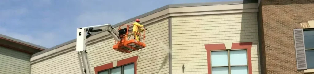 A worker in a yellow safety shirt and hard hat stands in an orange boom-lift basket, pressure-washing the upper exterior wall of a retail building with beige siding and red-trimmed windows.